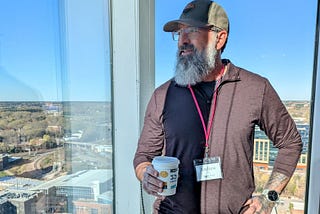 The author, a bearded man wearing glasses, a baseball cap, and a dark long-sleeve shirt, stands in front of a tall glass window overlooking a cityscape. He holds a coffee cup in one hand and wears a visible name badge that reads “Andrew.” A pink lanyard is around his neck, and he has visible tattoos on his forearm and a smartwatch on his wrist. The background shows a clear blue sky, buildings, and trees.