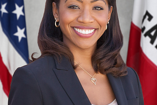 Malia Cohen, an African-American women, smiling broadly and warmly toward the camera. She is wearing a black suit jacket and white tank top. Behind her are the US flag and the California state flag.