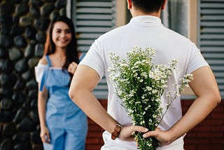 Man holding flowers behind his back while girlfirend looks on in anticipation.