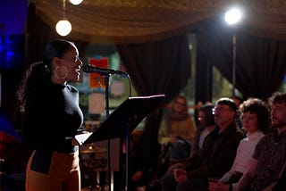 A photo of a journalist talking into a microphone to an audience of people.