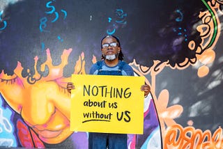 A Deaf Black man with a septum piercing and a black and white beard, wearing denim overalls and glasses, holds a hand-lettered sign that reads ‘NOTHING about us without US.’ A vibrant mural of a Black woman serves as the background.