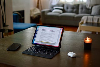 IPad Pro in magic keyboard on dining room table next to AirPods, iPhone, and lit candle.