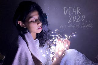 Sitting with fairy lights that are in a ball, wearing a scarf. Labeled, “Dear 2020…my deepest thoughts”.