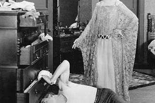 A man and woman in a messy room, and the man is trying to find something underneath the dresser.