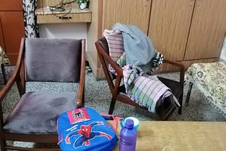 This is a sofa set alongwith blankets ,school bag ,water bottle and a cable wire in the background.