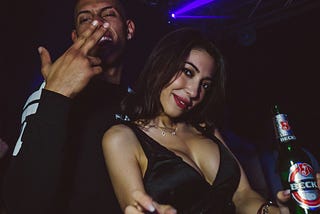 Man and woman in a party scene. Man grinning, making a hand sign, woman in a low cut dress, smiling while holding a beer.