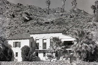 Black and white photograph of The Willows in Palm Springs. The Mediterranean-style villa sits at the base of a rocky hillside with the San Jacinto Mountains rising behind it. The white stucco exterior features arched windows, a tiled roof, and a small balcony, with palm trees and desert landscaping surrounding the residence.