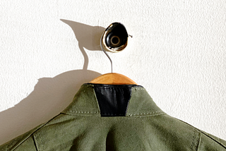 Photograph of the back of an army green jacket, hanging from a hanger. The collar has been sliced in half, and a black leather triangular patch has been sewn in to create a ‘collar gap’.