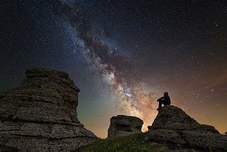 A person sits on rocks at night looking up at the Milky Way Galaxy.