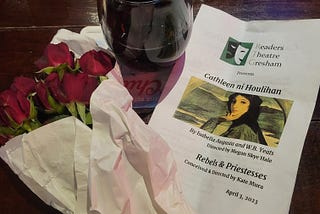 Small red roses wrapped in paper, a tumbler of red wine, and slightly crumpled program for Readers Theatre Gresham.