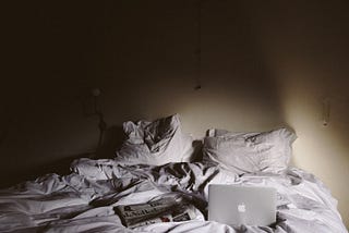 White crumped bed sheets in a dark room, with an open MacBook and a newspaper strewn across the bed
