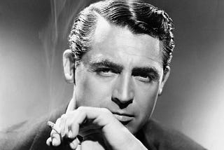 Classic film actor, Cary Grant poses with a cigarette in one hand. Smoke billows beside his face.