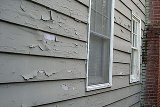 Gray wood siding with peeling paint and two white windows.