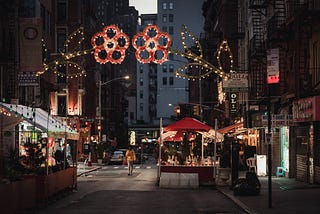 A street shot of a Little Italy in the States. Decorative streetlights in the shape of red flowers create an archway over the street. There are small food stalls slipping out of the sideway and onto the narrow city street.