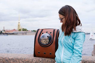 A lady standing close to a lake with a cat in a cat backpack.