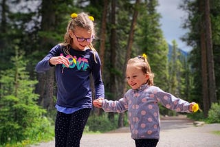 An older and younger girl holding hands as they walk down a dirt road flanked by forest