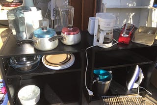 A close up shot of two small black side tables with household items such as saucepans, a coffee maker, a juicer, and iron, some baking ware. All this is on a front porch, ready to give away.