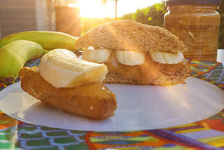 Two peanut butter and banana sandwiches on a plate, bananas, and a peanut butter jar. The sun is shining in the background.