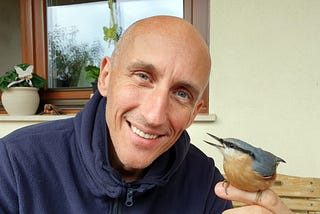 The author, József Manhertz, looks into the camera with a smile. He is bald and has blue eyes. A Eurasian nuthatch sits on the index finger of his left palm.