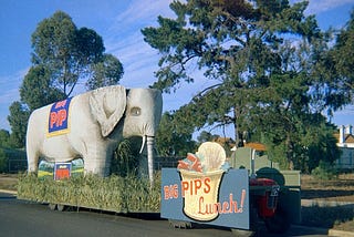 Life-sized elephant parade float “Big PIP” being towed by a tractor, hidden by advertising for “Pips” lunch.