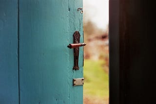 A green door with brown handle is slightly opened to a bright day outside.