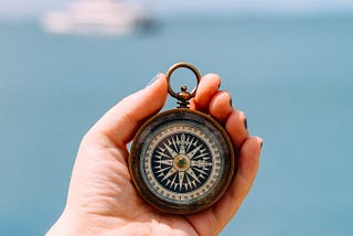 A hand holding up a nautical compass, in front of a large stretch of ocean. In the background is a ship.
