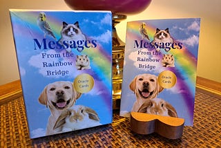 A box of oracle cards leans against a lamp on an end table with the guide booklet to its right. The oracle card box and booklet shows a blue sky with white cloud and a rainbow with pictures of a bird, cat, hamster, dog and rabbit. The words are: Messages From the Rainbow Bridge. Oracle Cards.