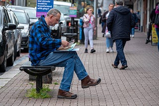 Rick, a middle aged white man, wearing a blue and black checked shirt and blue jeans, sitting on a black bench looking through an activity pack. People and signs on the High Street behind him.