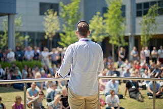 A man speaks to a crowd, who are seated on the ground in front of him