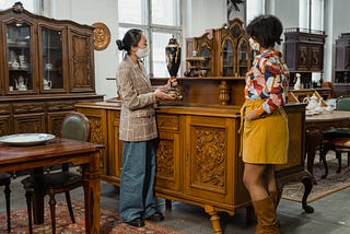 Two women standing in front of an antique side table wearing masks