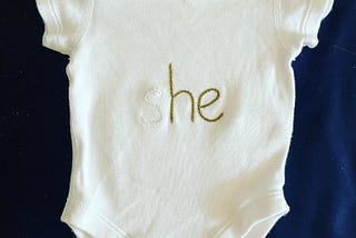 a white babygrow with the words “she” embroidered on it. The S is in white, the HE is in green. This represents how I feel my daughter is rendered invisible by the linguistic patriarchy in many parenting books and articles.