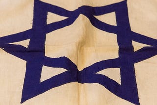 A hand-sewn flag, feature a blue Star of David at the center and two horizontal blue cloth stripes near the top and bottom.