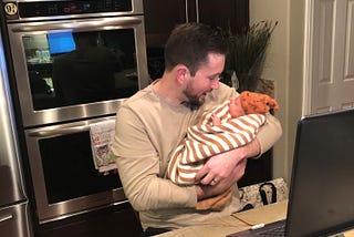 Brett holding baby Ezra in the kitchen while working on a laptop. Brett is looking down and smiling at Ezra. A Platform 9 and 3/4 magnet can be spotted in the background.