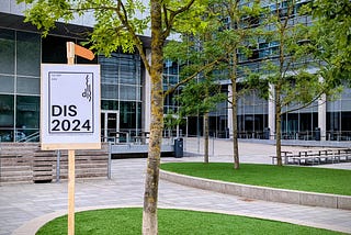 Photo of the conference entrance, showing a DIS 2024 sign, some trees, and the building and entrance.