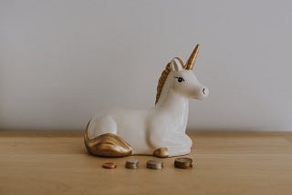 Ceramic white unicorn with a gold horn sitting in front of stacks of coins.