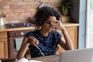 Young woman of color presses a finger to the bridge of her nose in clear frustration as she sits in front of a laptop.