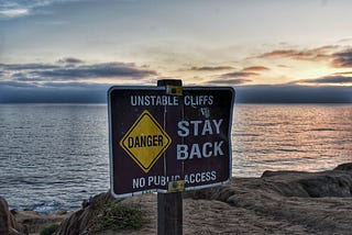Signage upon a body of water that reads “Unstable Cliffs Stay Back”