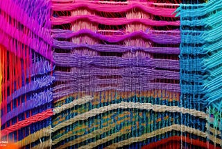 Adobe Firefly generated image with the prompt: Generate an image of a loom weaving threads into a patterned tapestry, where the threads are lines of code, showing how Generative AI weaves together different threads of data to form a coherent output. Use a palette of vibrant colors to represent the variety of data.