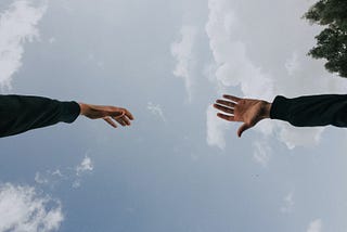 A photograph of two hands reaching for each other, with the sky in the background