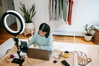 Woman working on a laptop with a notebook beside her while a ring light and smartphone recording setup dominate the foreground, illustrating the overlap between writing and online content creation.
