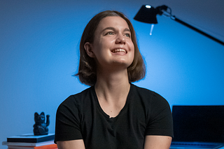 The author in front of her desk, in the background there is a monitor, desk lamp, and pile of notebooks with a simple black decoration.