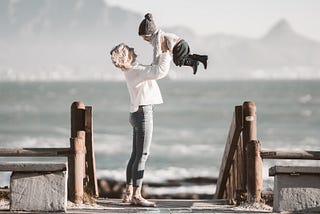 blonde woman is throwing toddler boy up in the air next to a lake and mountains