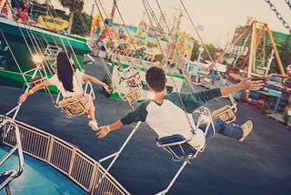 Two people seen from behind ride on a fairground swing with their arms out.