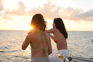 Two young women who seem to be friends at a body of water with drinks. They are watching the sunset together, enjoying life.