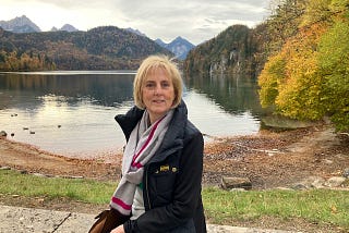 Susan faces the camera, she’s sat in front of a beautiful lake and is wearing a scarf and coat