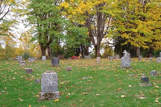 Photo of slanted cemetery with decaying old graves and autumnal leaves on the ground, trees in distance.