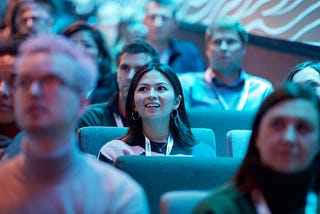 The image shows an audience seated in what appears to be a conference or event setting. The focus is on a young woman in the center, wearing a lanyard and looking engaged, with a bright and excited expression. She is surrounded by other attendees, some of whom are slightly out of focus. The lighting in the room is predominantly blue, creating a cool-toned ambiance. The background suggests that everyone is attentively watching a presentation or speaker. The setting appears modern and professional