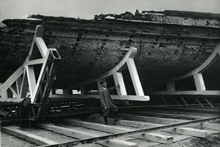 Black and white photograph of a man standing in front of a giant wooden hull