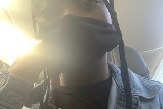 A selfie of me, sitting in a plane. Half of my face is covered by a mask, and my hair is styled in box braids. My facial expression is bleak.