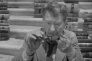 Screenshot of 1959’s Twilight Zone episode "Time Enough at Last", showing Henry Bemis holding his broken glasses amid books.